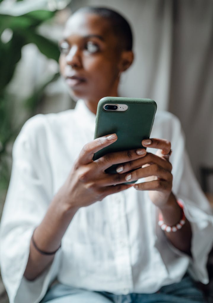 Calm pensive bald African American female in white casual blouse sitting on chair near big plant and browsing modern mobile phone while looking away thoughtfully